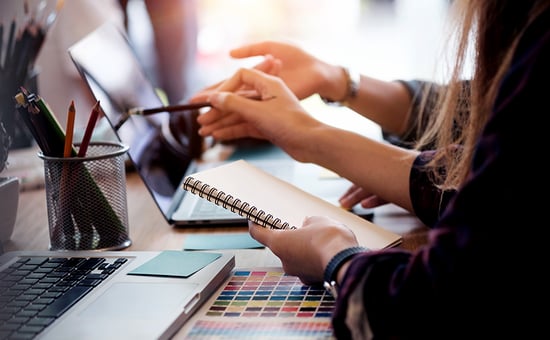 Two people's arms, hands pointing at a laptop and holding a notebook